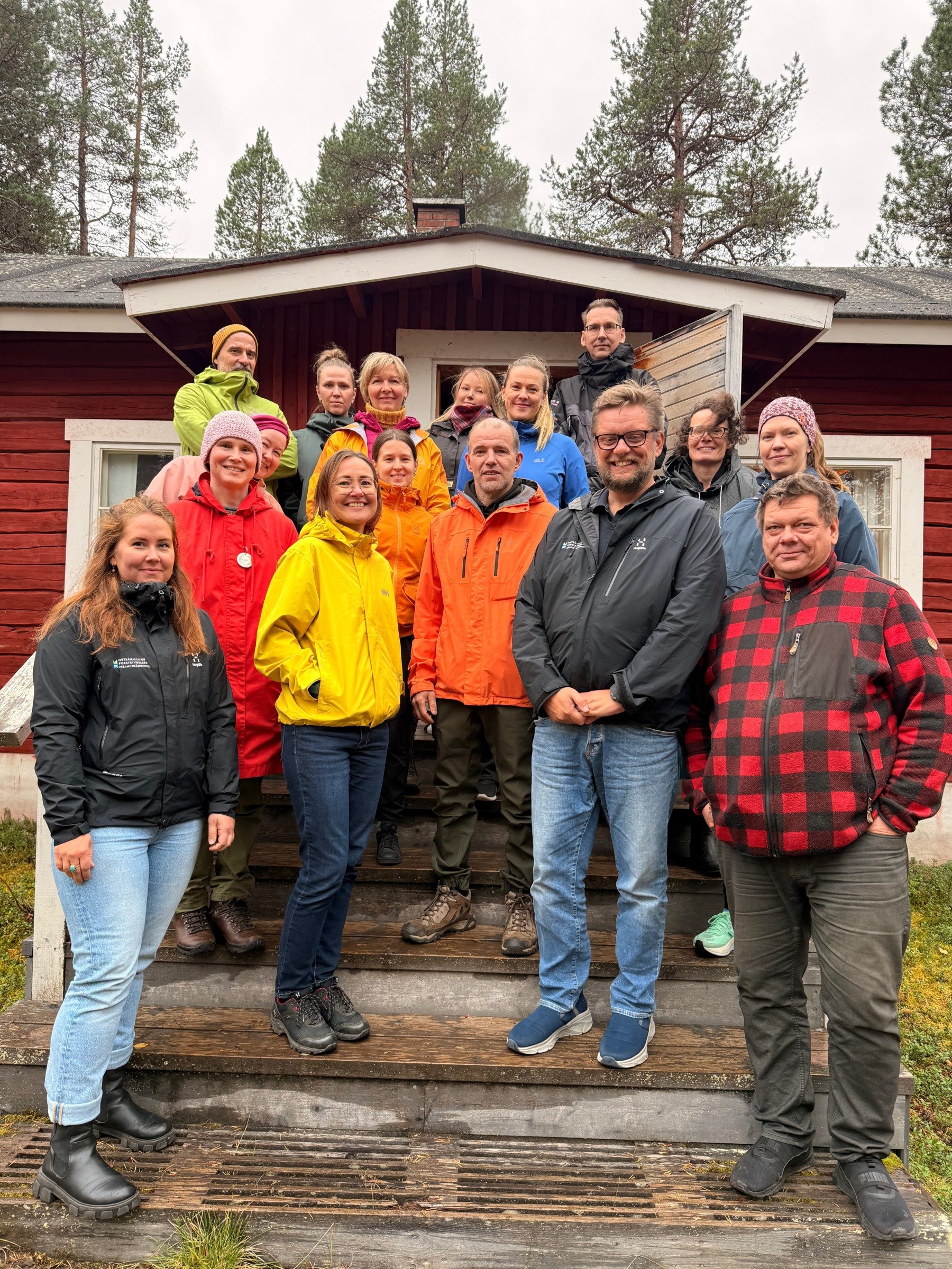 Ryhmä ihmisiä seisoo ulkona puutalon portailla. He katsovat kameraan ja hymyilevät. Taustalla näkyy metsää ja pilvinen taivas.